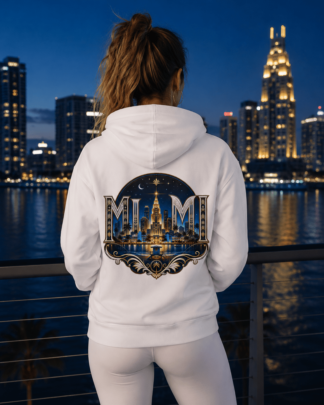 A woman viewed from behind wearing a white hoodie with a large Miami skyline graphic on the back, standing on a balcony with a city skyline at dusk in the background.