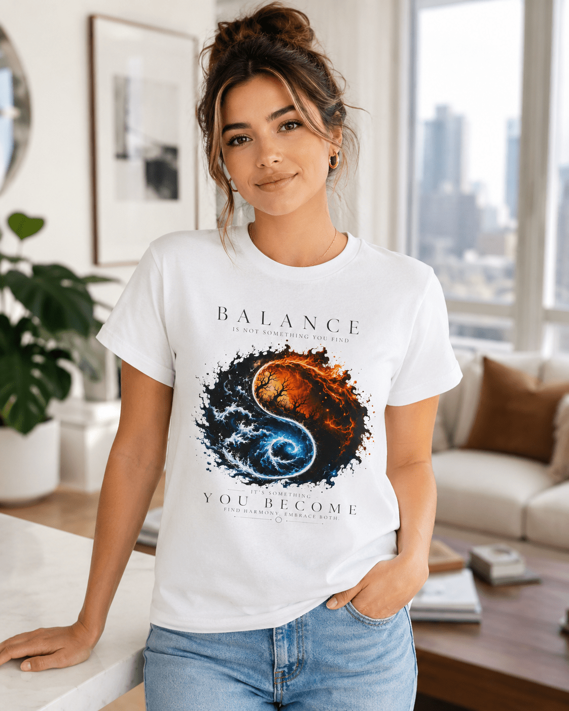 A woman wearing a white graphic t-shirt with a yin-yang style design featuring a fiery orange and blue cosmic print, paired with light blue jeans in a bright, modern living room setting.