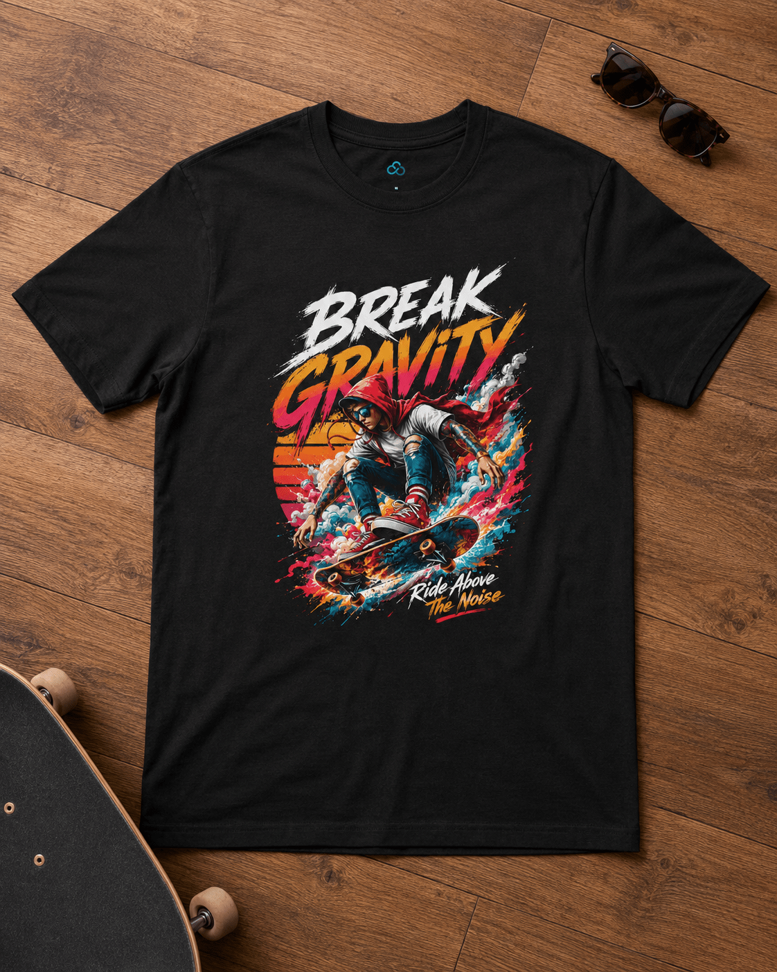 A black short-sleeve graphic T-shirt laid flat on a wooden floor, featuring a colorful skateboarding design with the words 'BREAK GRAVITY' and 'Kids Have More Fun'. Sunglasses and parts of a skateboard are placed nearby as props.