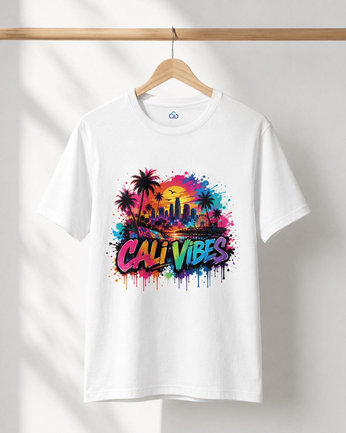 A white short-sleeve crewneck t-shirt hanging on a wooden hanger against a white wall. The shirt features a colorful graphic print on the front with palm trees, a city skyline, a sunset, and the text 'CALI VIBES' in a graffiti-style font.