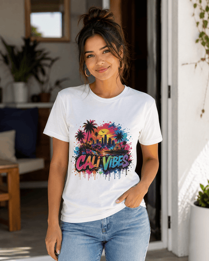 A woman wearing a white crewneck t-shirt with a colorful 'CALI VIBES' graphic print featuring palm trees and a sunset skyline, paired with light blue jeans, standing in a bright, modern indoor setting.