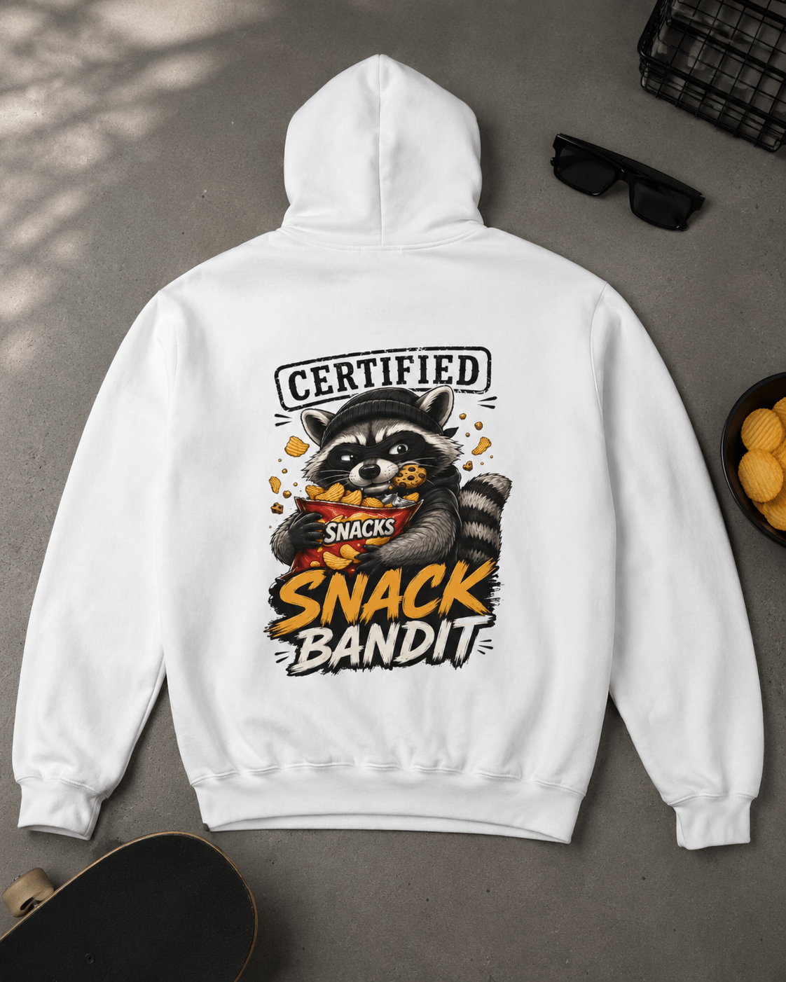 Flat lay view of a white hoodie displayed on a concrete floor with a skateboard, sunglasses, and a bowl of chips, featuring a back graphic of a raccoon eating chips with text reading 'Certified Snack Bandit'.