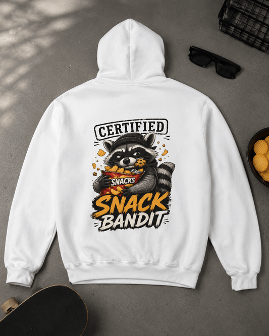 Flat lay view of a white hoodie displayed on a concrete floor with a skateboard, sunglasses, and a bowl of chips, featuring a back graphic of a raccoon eating chips with text reading 'Certified Snack Bandit'.