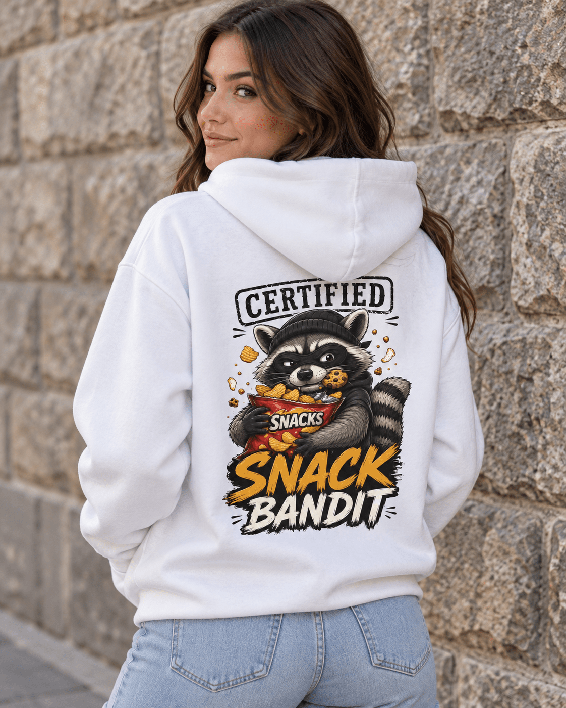 A woman wearing a white hoodie with a large raccoon snack graphic and the text 'CERTIFIED SNACK BANDIT' on the back, standing against a stone wall.