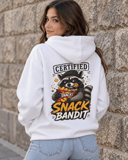 A woman wearing a white hoodie with a large raccoon snack graphic and the text 'CERTIFIED SNACK BANDIT' on the back, standing against a stone wall.
