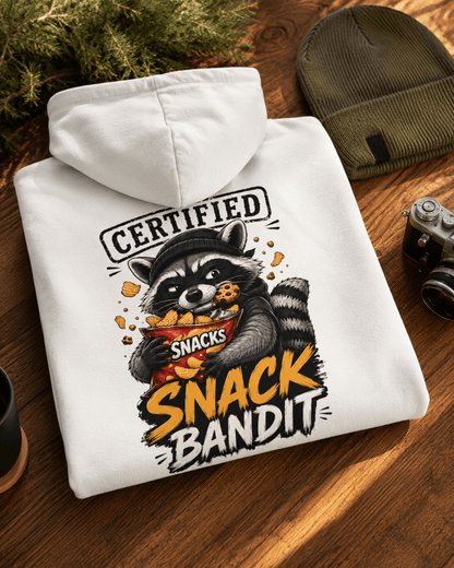 Folded white hoodie on a wooden table beside a vintage camera and green beanie, showcasing a back print of a raccoon wearing a mask and holding a chip bag with 'Certified Snack Bandit' text.