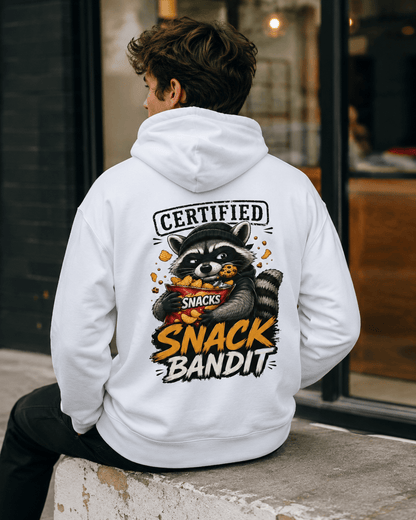 Man seated outdoors turned away from the camera wearing a white hoodie, showcasing a back graphic of a raccoon eating chips and a cookie with text reading 'Certified Snack Bandit'.