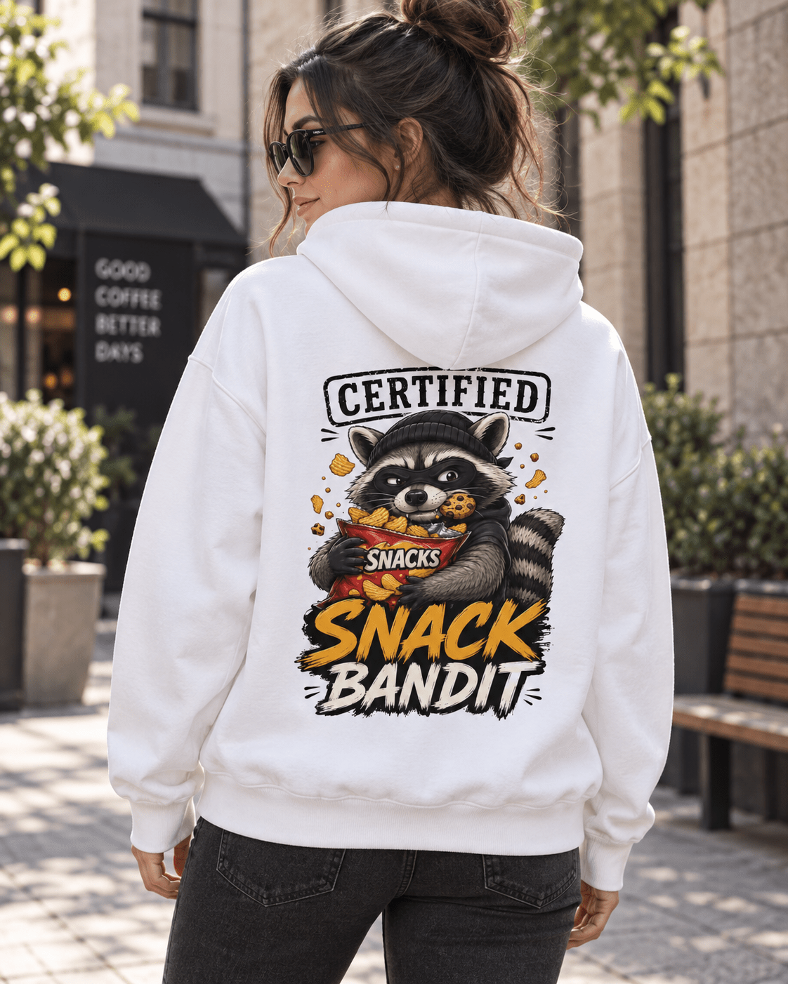 Back view of a woman walking on a city sidewalk wearing a white hoodie, featuring a graphic of a raccoon with a cookie in its mouth holding a red snack bag and text reading 'Certified Snack Bandit'.