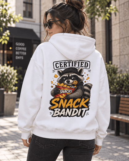 Back view of a woman walking on a city sidewalk wearing a white hoodie, featuring a graphic of a raccoon with a cookie in its mouth holding a red snack bag and text reading 'Certified Snack Bandit'.