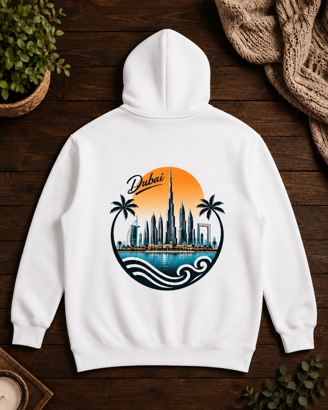 A white pullover hoodie shown from the back, laid flat on a dark wood surface with a colorful Dubai cityscape graphic printed in the center. The design includes palm trees, the Burj Khalifa skyline, waves, and an orange sun with the word 'Dubai' in script.