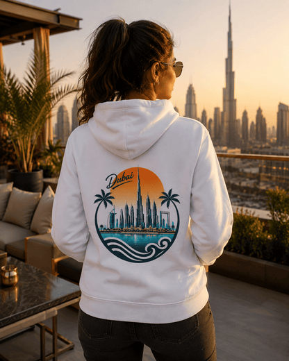 A woman viewed from behind wearing a white hoodie with a large graphic print on the back featuring the word 'Dubai', palm trees, the Burj Khalifa skyline, and waves. She is standing on a balcony with a sunset view of Dubai's skyline in the background.