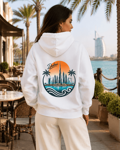 A woman viewed from behind wearing a white hoodie with a large circular Dubai cityscape graphic on the back, featuring palm trees, Burj Khalifa, waves, and an orange sun, set against an outdoor waterfront patio background.