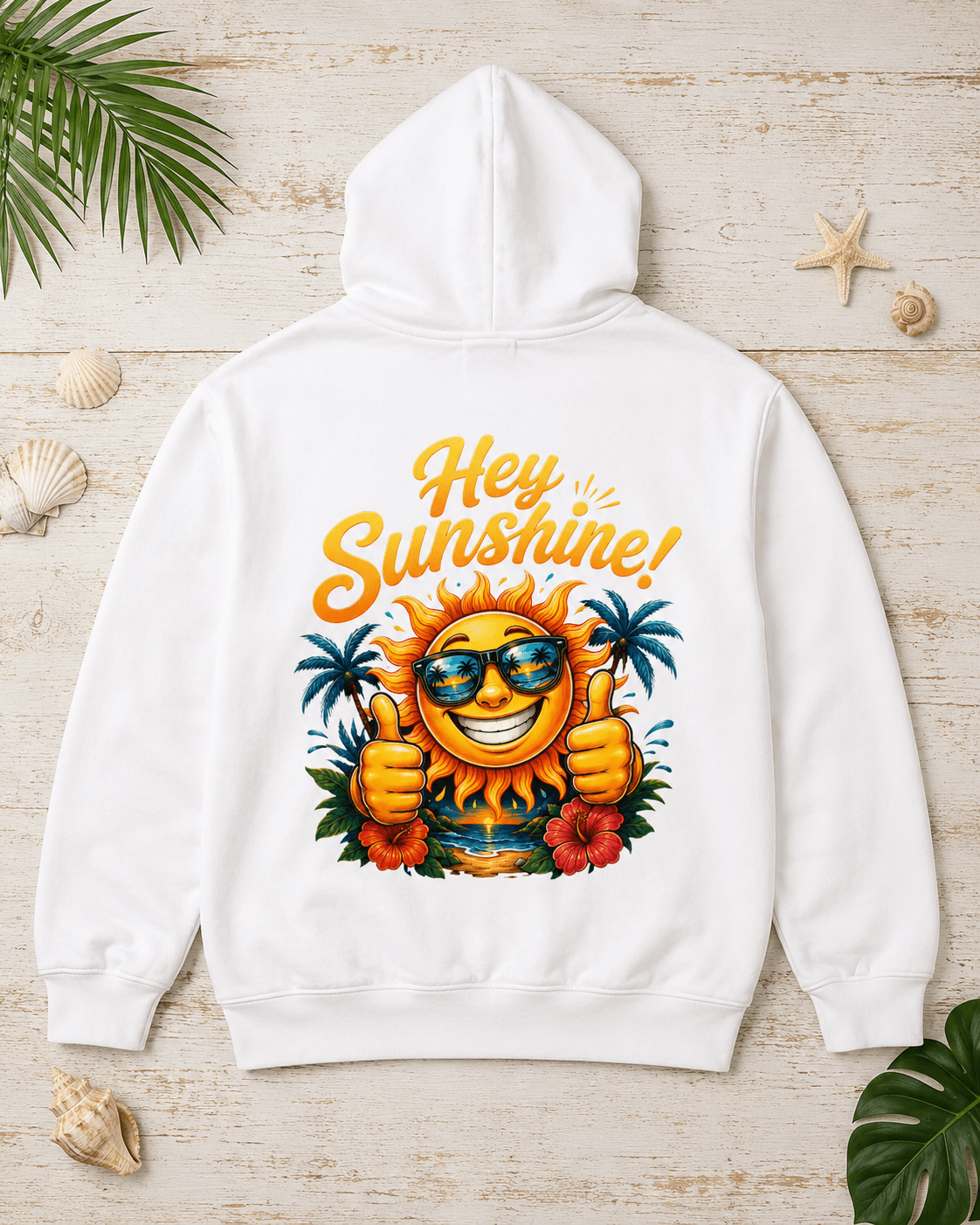 A white hooded sweatshirt laid flat on a light wood surface, decorated with seashells and green leaves. The back of the hoodie features a colorful graphic of a smiling sun wearing sunglasses, giving two thumbs up, with palm trees and hibiscus flowers, and the text 'Hey Sunshine!' in yellow-orange lettering.