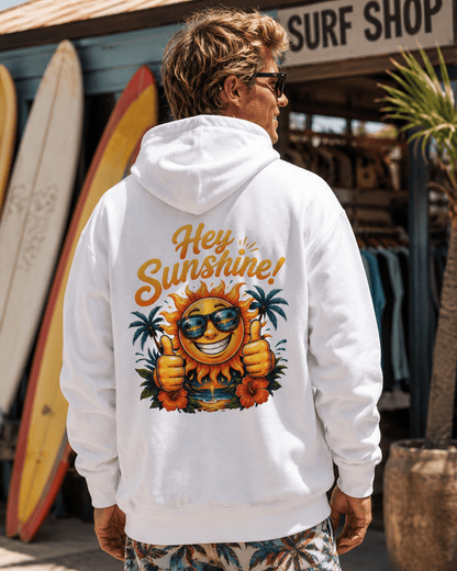 A man wearing a white hoodie with a colorful graphic on the back that reads 'Hey Sunshine!' and shows a smiling sun, sunglasses, palm trees, and flowers. He is standing in front of a surf shop with surfboards visible in the background.