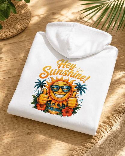 A folded white hooded sweatshirt with a colorful graphic of a smiling sun wearing sunglasses and giving two thumbs up, surrounded by palm trees and flowers, with the text 'Hey Sunshine!' above it. The hoodie is displayed on a wooden surface with a woven mat and plant leaves in the background.
