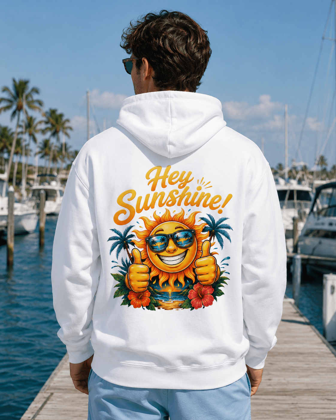 A man viewed from behind wearing a white hoodie with a large colorful graphic on the back that reads 'Hey Sunshine!' with a smiling sun, sunglasses, palm trees, and flowers, standing on a wooden dock by the water.