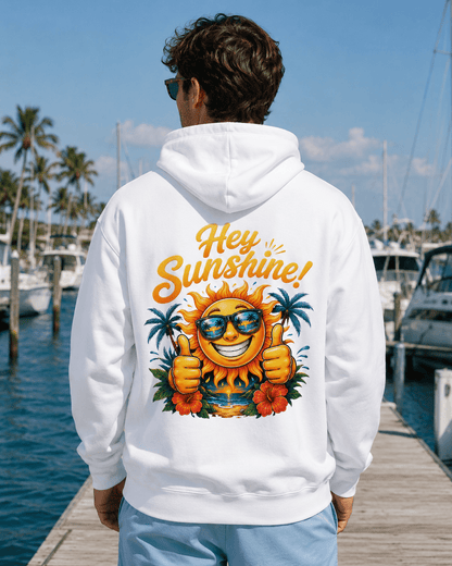 A man viewed from behind wearing a white hoodie with a large colorful graphic on the back that reads 'Hey Sunshine!' with a smiling sun, sunglasses, palm trees, and flowers, standing on a wooden dock by the water.