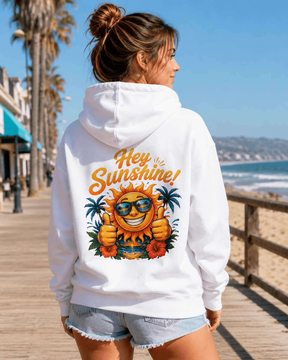 A woman viewed from behind wearing a white hoodie with a large colorful graphic on the back that reads 'Hey Sunshine!' with a smiling sun, sunglasses, palm trees, and flowers. The photo is taken on a sunny boardwalk near the beach.