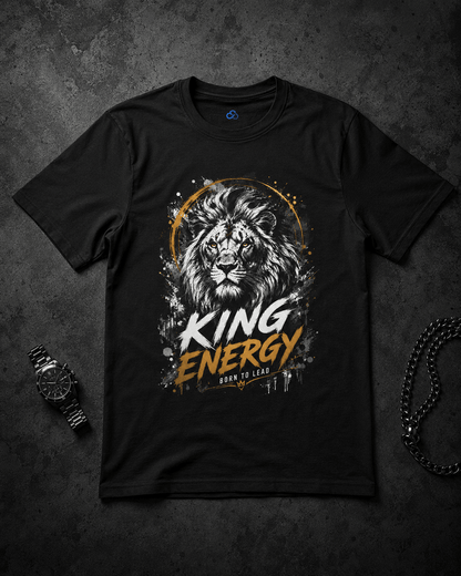 A black short-sleeve graphic t-shirt laid flat on a dark textured background, featuring a lion illustration with the words 'KING ENERGY' in white and orange. A watch and a chain are placed beside the shirt as styling props.