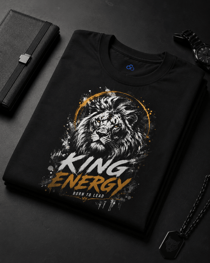 A folded black crewneck t-shirt with a graphic print of a lion's face, gold splatters, and the text 'KING ENERGY' and 'BORN TO LEAD', styled on a dark surface with a notebook, watch, and dog tags nearby.