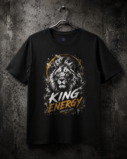 A black short-sleeve t-shirt hanging on a metal hanger against a dark textured wall. The shirt features a graphic print of a lion's face inside a gold circular frame, with the words 'KING ENERGY' in white and gold distressed lettering, and 'BORN TO LEAD' below it.