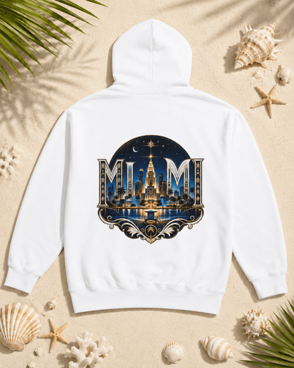 A white hooded sweatshirt laid flat on a sandy surface with seashells and palm leaves around it. The back of the hoodie has a large circular graphic featuring the word 'MUM' in ornate lettering over a nighttime city skyline with a glowing tower and blue sky.