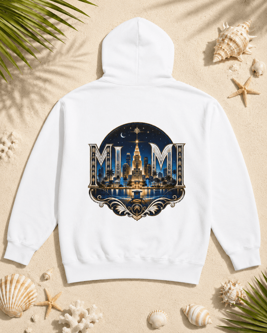 A white hooded sweatshirt laid flat on a sandy surface with seashells and palm leaves around it. The back of the hoodie has a large circular graphic featuring the word 'MUM' in ornate lettering over a nighttime city skyline with a glowing tower and blue sky.