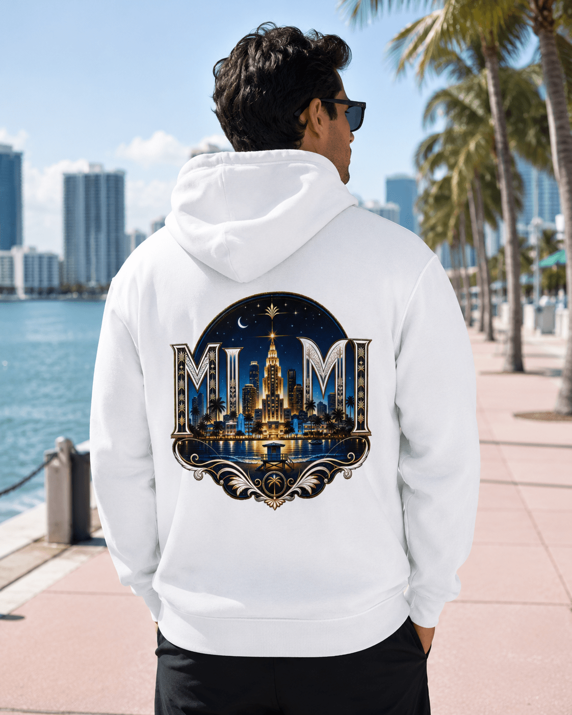 A man wearing a white hoodie with a large 'MUM' Miami skyline graphic on the back, standing on a waterfront walkway with palm trees and a city skyline in the background.