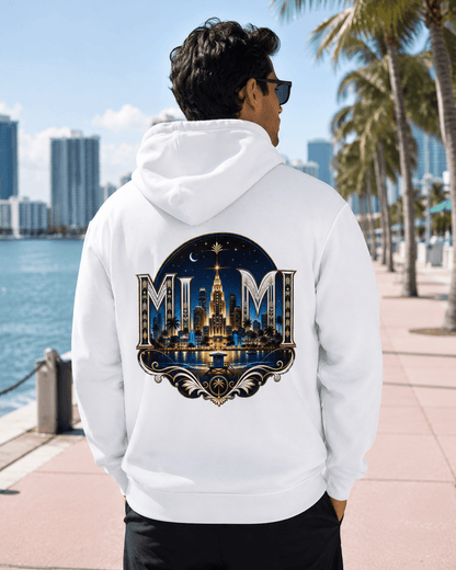 A man wearing a white hoodie with a large 'MUM' Miami skyline graphic on the back, standing on a waterfront walkway with palm trees and a city skyline in the background.