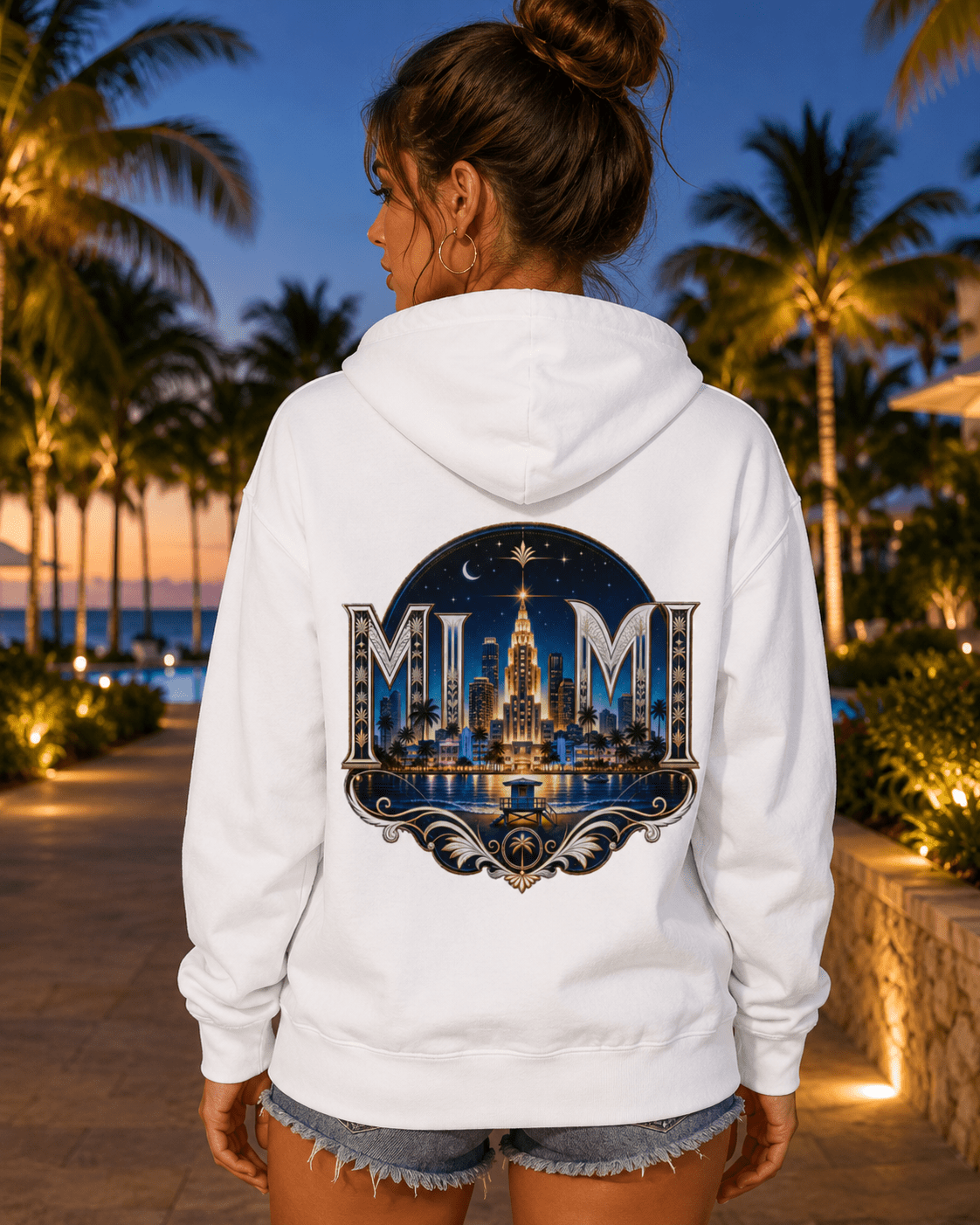 A woman viewed from behind wearing a white pullover hoodie with a large Miami skyline graphic on the back, set against a tropical beach path at dusk.