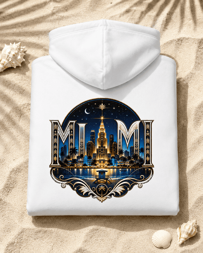 A white hooded sweatshirt laid flat on sandy ground with seashells nearby. The front has a large decorative graphic featuring the letters 'MIM' and a nighttime city skyline with a tall illuminated building, stars, moon, and water reflection.