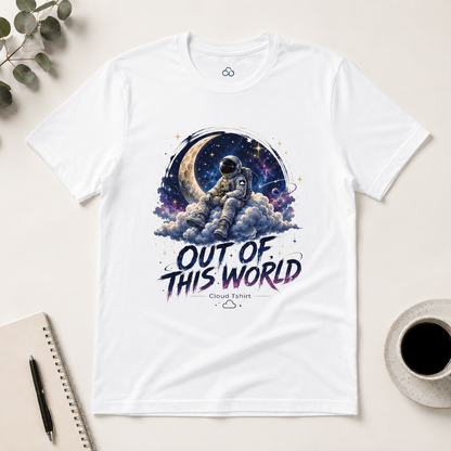 A white short-sleeve graphic T-shirt laid flat on a light beige surface, featuring an astronaut sitting on clouds with a crescent moon and starry sky behind them, and the text 'OUT OF THIS WORLD' with 'Cloud T-Shirt' underneath. A notebook, pen, eucalyptus leaves, and a cup of coffee are arranged around it as styled props.