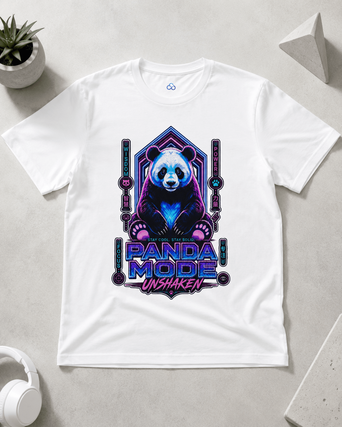 A white short-sleeve crewneck t-shirt laid flat on a light gray surface, featuring a colorful graphic of a panda bear in a neon purple and blue geometric frame with the text 'PANDA MODE UNSHAKEN'. A small potted plant, white headphones, and geometric props are visible around the shirt.