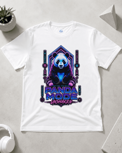 A white short-sleeve crewneck t-shirt laid flat on a light gray surface, featuring a colorful graphic of a panda bear in a neon purple and blue geometric frame with the text 'PANDA MODE UNSHAKEN'. A small potted plant, white headphones, and geometric props are visible around the shirt.