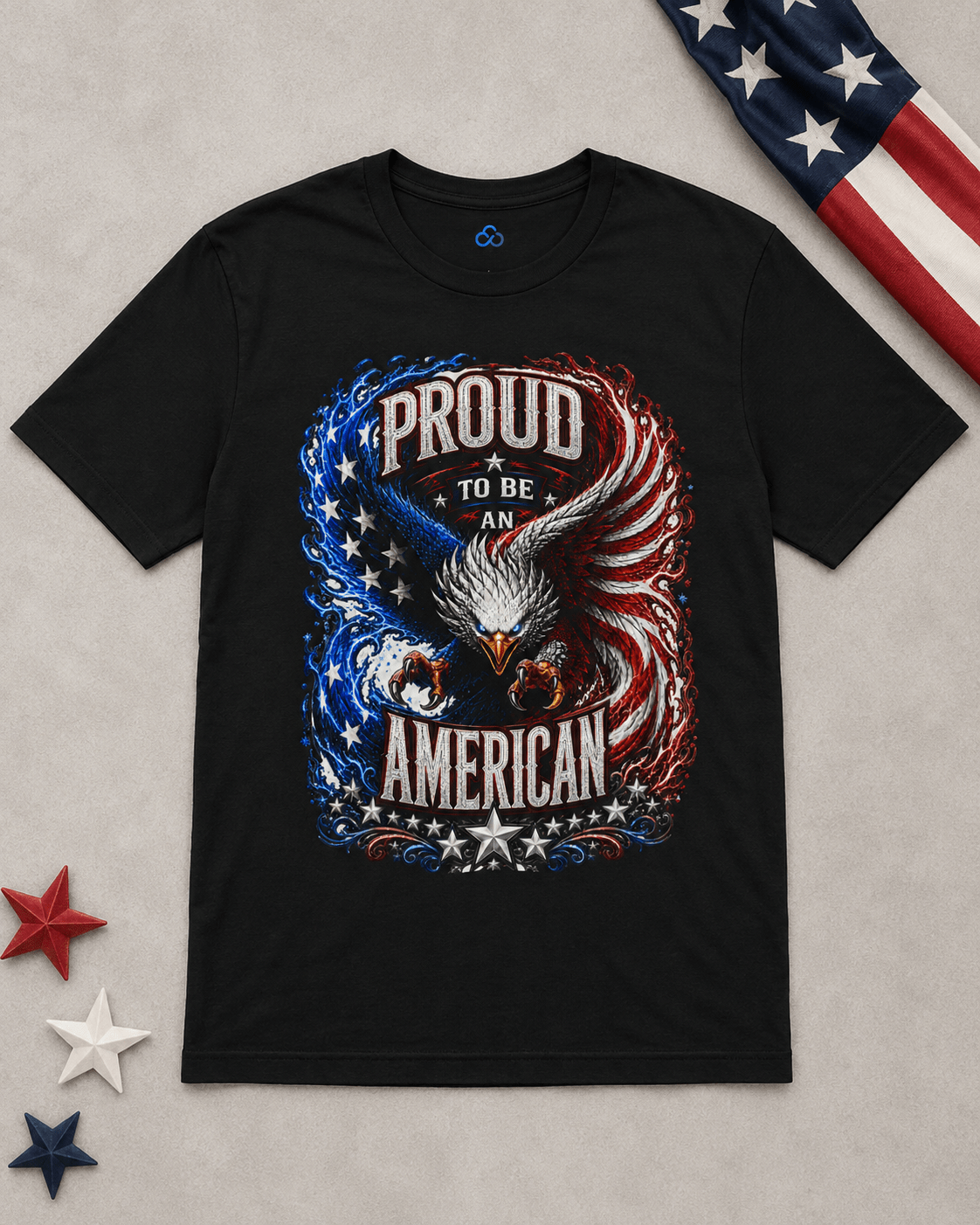 A black short-sleeve graphic T-shirt laid flat on a light gray surface, featuring a large patriotic eagle design with the words 'PROUD TO BE AN AMERICAN' in white, red, and blue. Decorative American flag elements and star cutouts appear around the shirt.