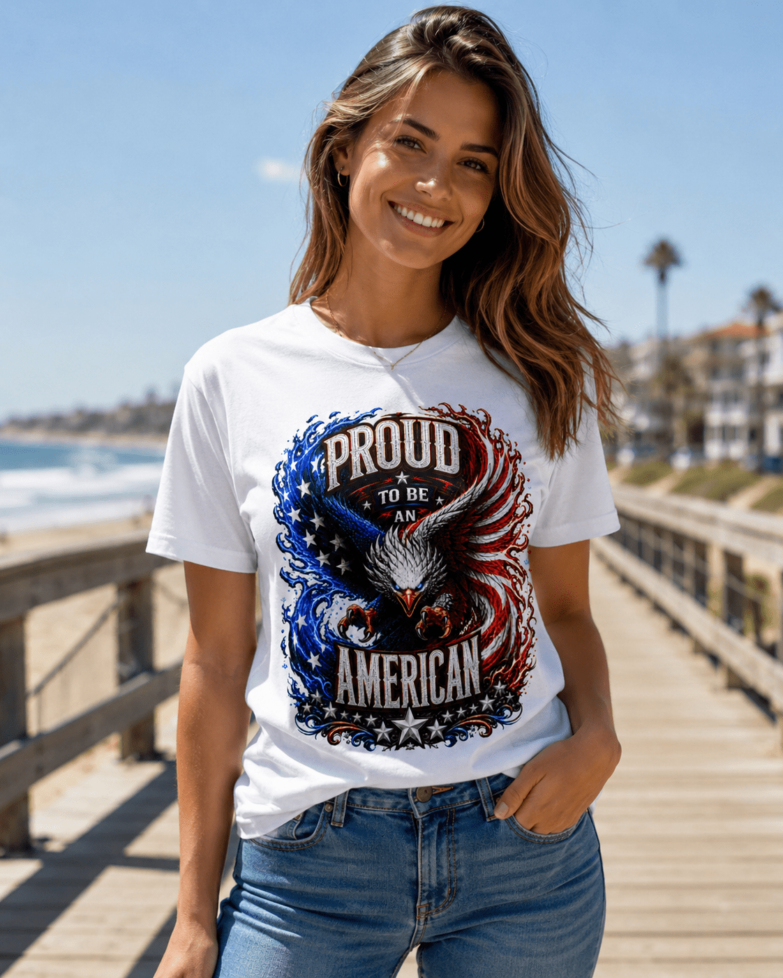 A woman wearing a white graphic t-shirt with a patriotic eagle design that reads 'PROUD TO BE AMERICAN' in red, white, and blue, standing on a wooden boardwalk by the beach.