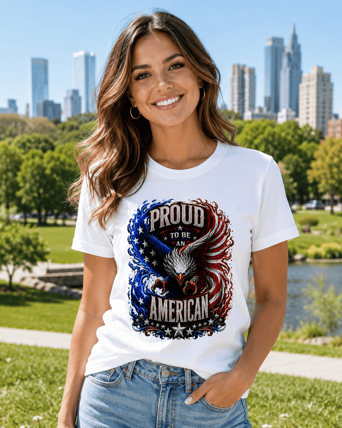 A woman wearing a white graphic t-shirt with a patriotic eagle design that reads 'PROUD TO BE AN AMERICAN' in red, white, and blue, set against a sunny park background with a city skyline.