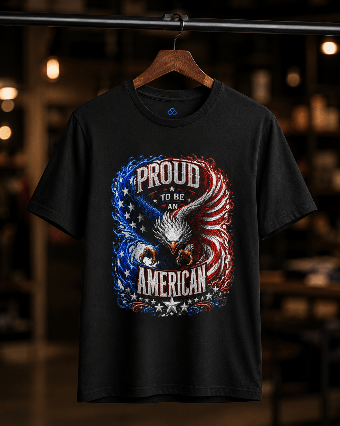 A black short-sleeve graphic t-shirt hanging on a wooden hanger against a blurred warm indoor background. The shirt features a large patriotic eagle graphic with the text 'PROUD TO BE AN AMERICAN' in distressed white lettering, with blue and red flag-inspired swirls and stars.