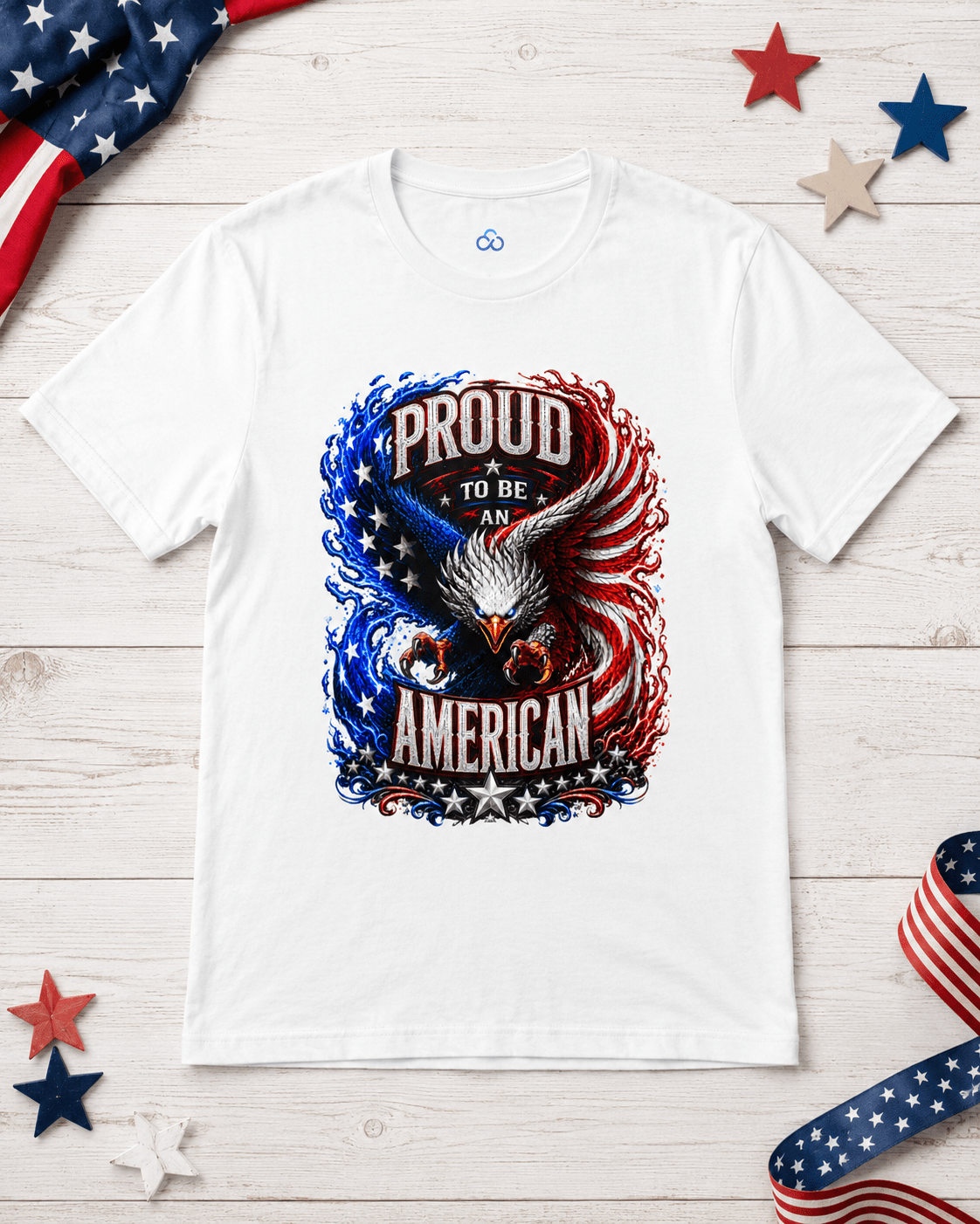 A white short-sleeve t-shirt laid flat on a light wood surface, featuring a patriotic graphic of an eagle and the words 'PROUD TO BE AMERICAN' in red, white, and blue. The shirt is styled with a US flag, red and blue stars, and a striped ribbon.