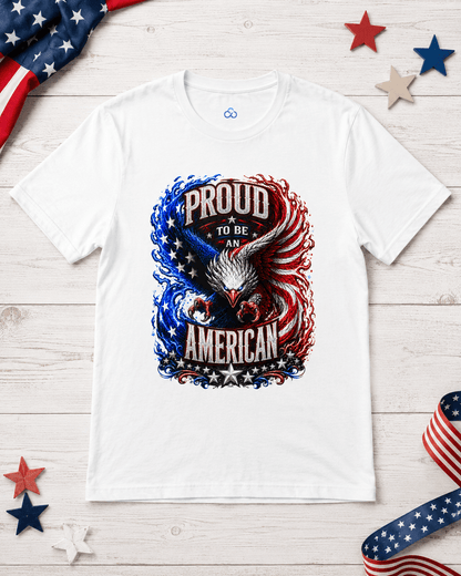A white short-sleeve t-shirt laid flat on a light wood surface, featuring a patriotic graphic of an eagle and the words 'PROUD TO BE AMERICAN' in red, white, and blue. The shirt is styled with a US flag, red and blue stars, and a striped ribbon.