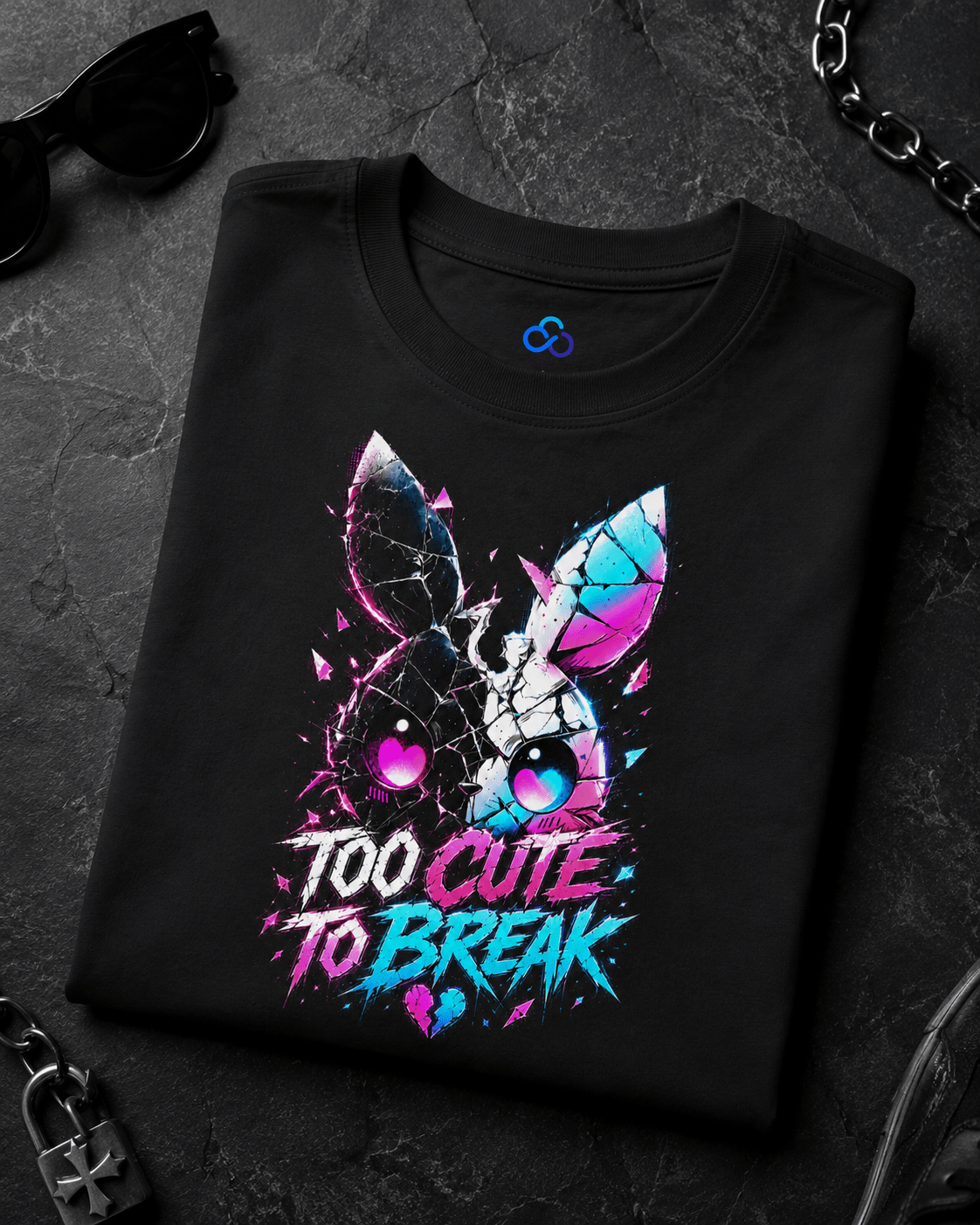 A black folded graphic T-shirt displayed on a dark textured surface with sunglasses, a chain, and a padlock as styled props. The shirt features a colorful bunny face with cracked glass details and the text 'TOO CUTE TO BREAK' in neon pink, blue, and purple.